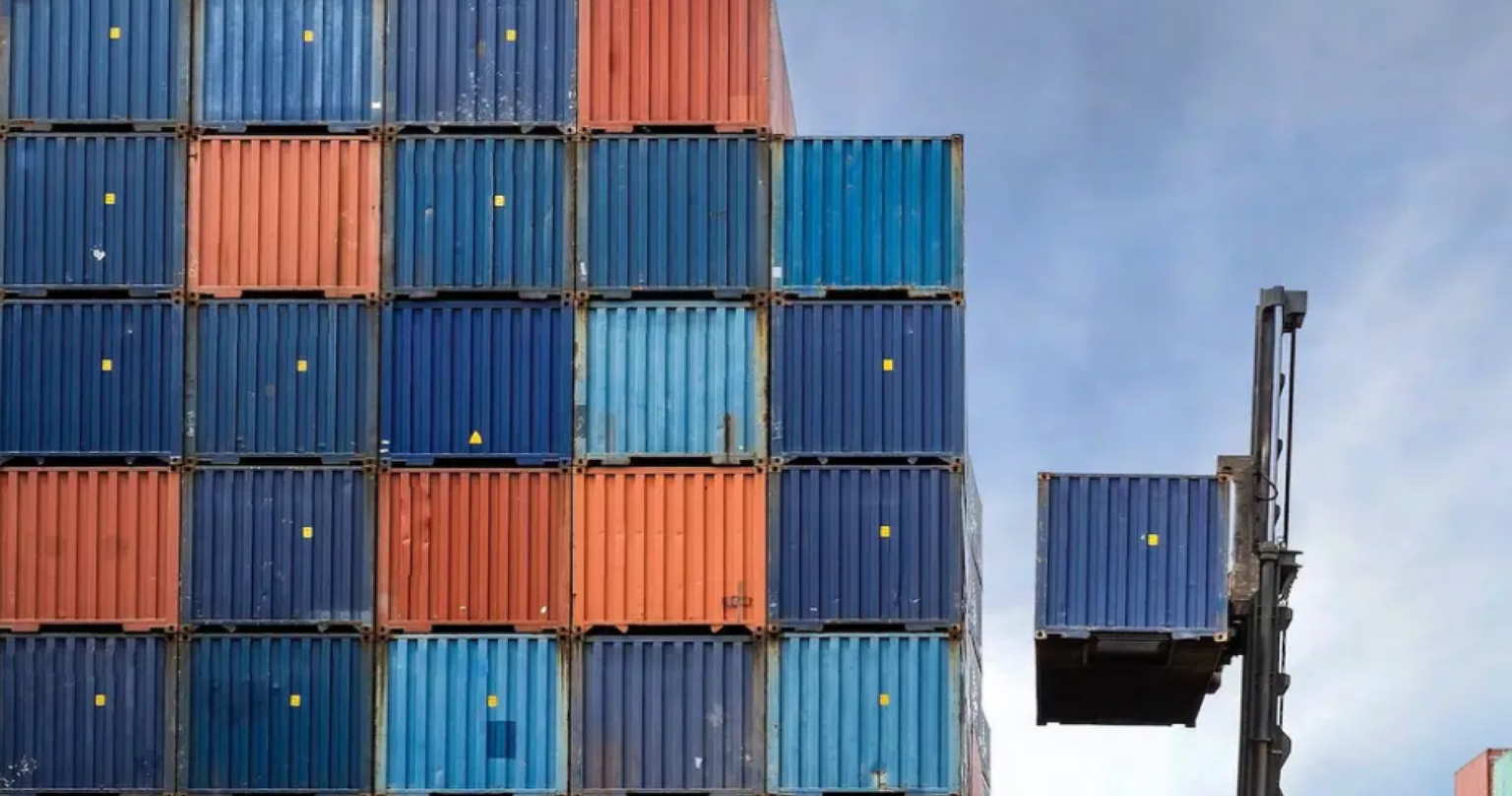 Blue, red, and orange shipping containers stacked in a block by a crane on a cloudy day.